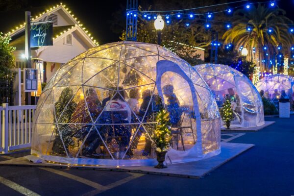People dine inside clear geodesic domes decorated with string lights and small Christmas trees, creating a cozy, festive outdoor atmosphere at night.
