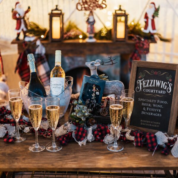 A festive holiday table with champagne glasses, bottles on ice, a reindeer figurine, and a Fezziwigs Courtyard sign offering food and drinks. Holiday decorations and a cozy fireplace are in the background.