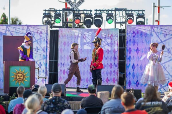 A group of actors in colorful costumes perform on an outdoor stage before an audience. The background features purple diamond patterns, and stage lights hang overhead. One actor wears a tall red hat, another holds a hand mirror.