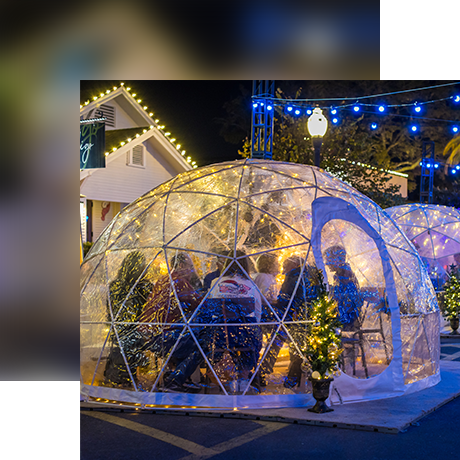 People sit inside a transparent, dome-shaped igloo decorated with string lights at night, surrounded by small lit trees and festive buildings in the background.