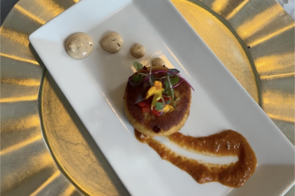 A gourmet appetizer on a white rectangular plate features a round patty topped with microgreens and edible flowers, a smear of orange sauce, and three dollops of beige sauce, all on a gold charger plate.