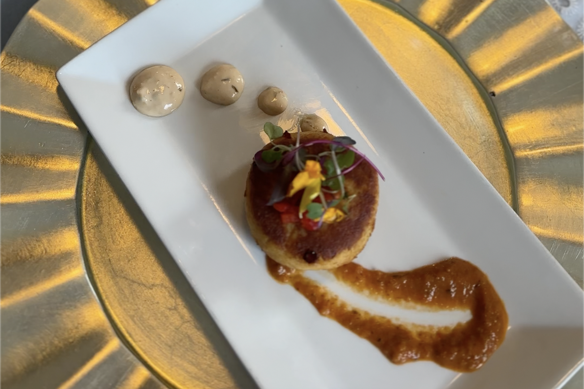 A gourmet appetizer on a white rectangular plate features a round patty topped with microgreens and edible flowers, a smear of orange sauce, and three dollops of beige sauce, all on a gold charger plate.