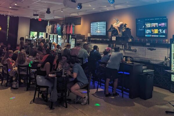 A lively bar scene with people sitting at tables and the bar, chatting and eating. TV screens display menus and images behind the bar, while shelves hold bottles and décor. The atmosphere is casual and social.