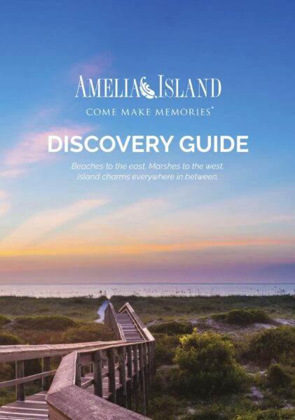 A wooden boardwalk leads over green dunes to a sandy beach at sunset, with pastel skies. Text reads: Amelia Island Discovery Guide. Beaches to the east. Marshes to the west. Island charms everywhere in between.