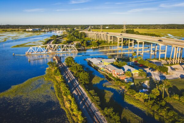 Aerial view of a river with a railroad drawbridge, a highway overpass, cars, green marshland, and buildings along the shoreline under a clear blue sky.