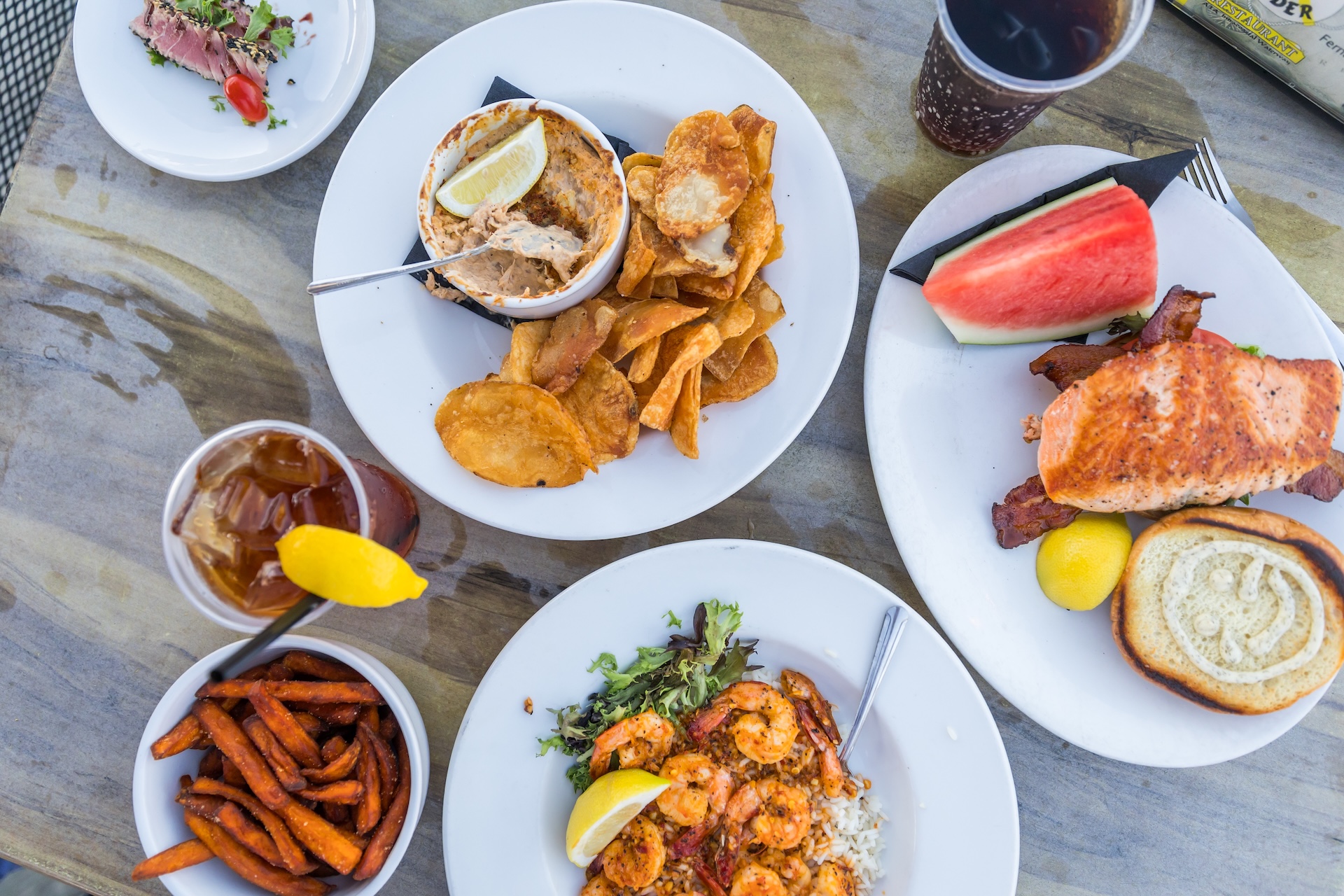 Overhead view of a restaurant table featuring fish dip with chips, a salmon sandwich with watermelon, shrimp over rice, sweet potato fries, and two drinks.