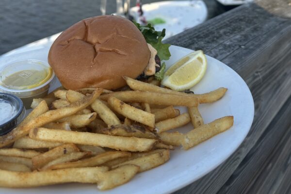 A plate with a sandwich bun, lettuce, a lemon wedge, a serving of French fries, and small sauce containers sits on a wooden surface by the water.