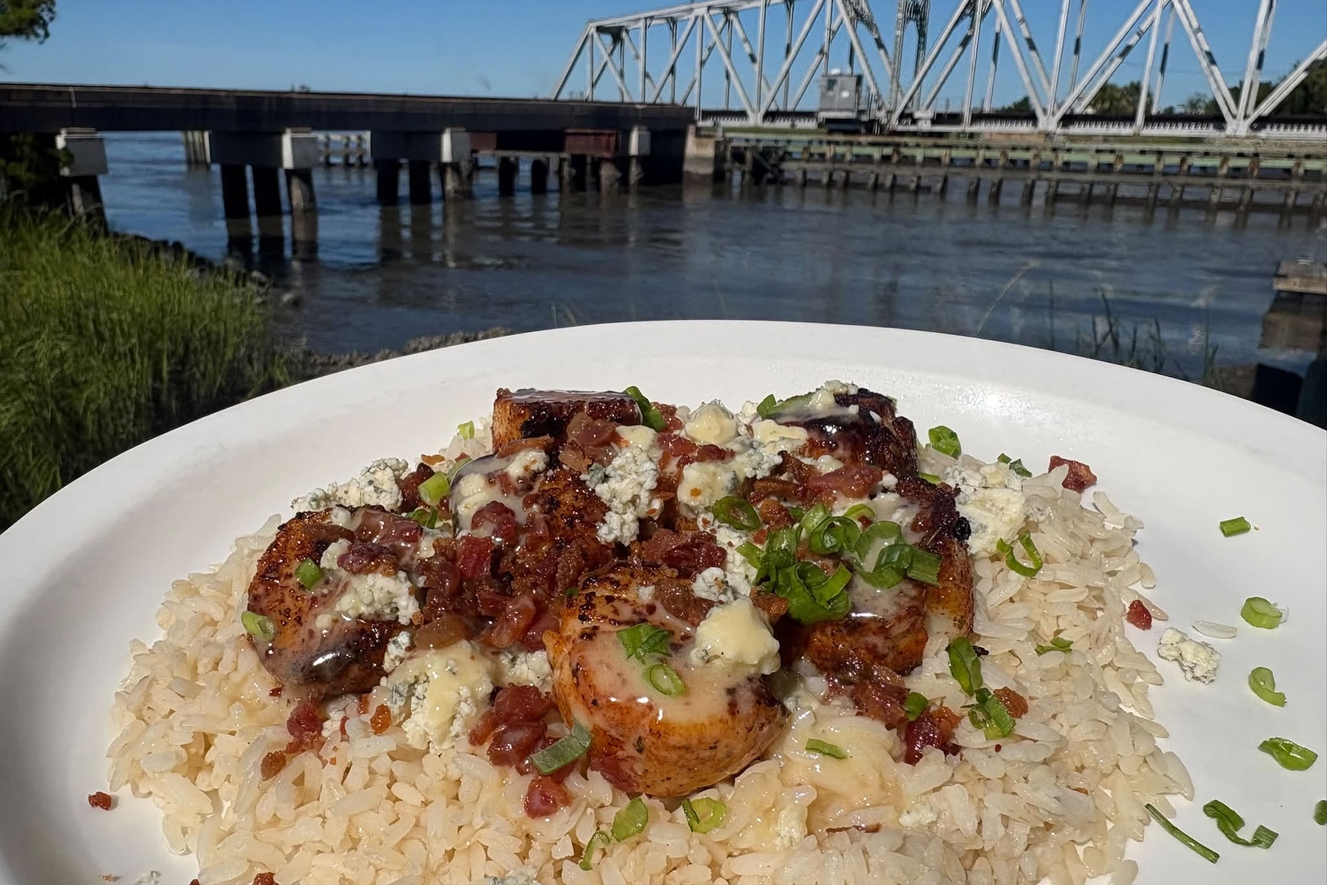 A white plate of rice topped with seasoned shrimp, crumbled cheese, bacon bits, and green onions, set outdoors by a river with a metal truss bridge in the background under a blue sky.