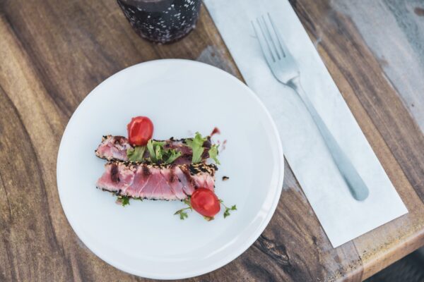 A white plate with two slices of seared tuna topped with greens and cherry tomato halves, placed on a wooden table next to a fork and a drink in a glass.