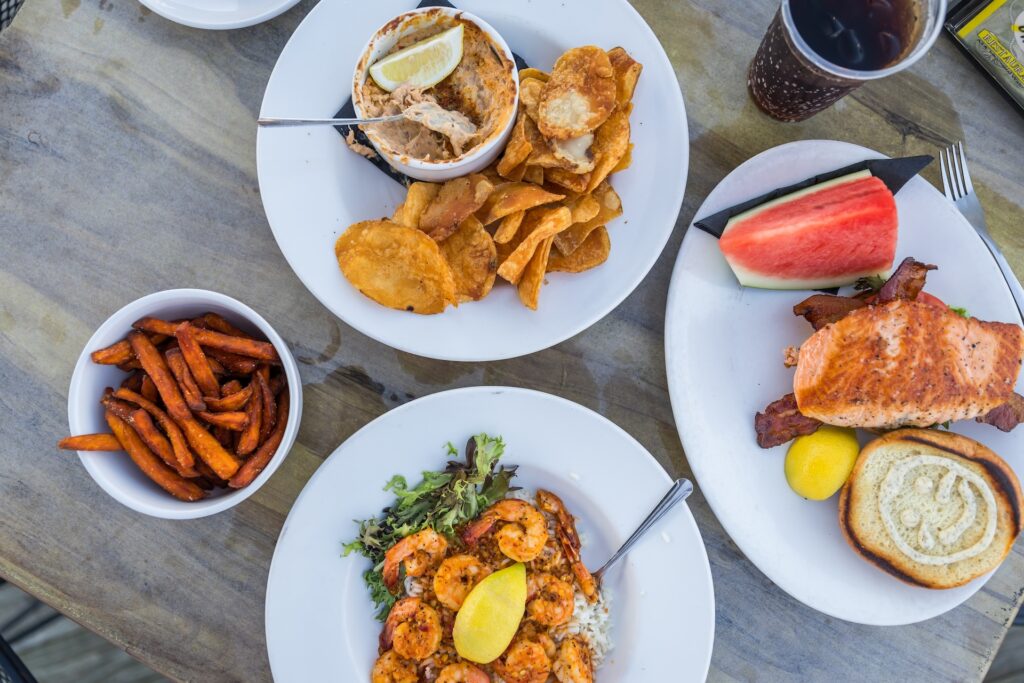 Four plates of food on a wooden table, featuring shrimp with rice and greens, sweet potato fries, a sandwich with bacon and watermelon, and a bowl of dip with thick-cut chips. There is also a drink with ice.
