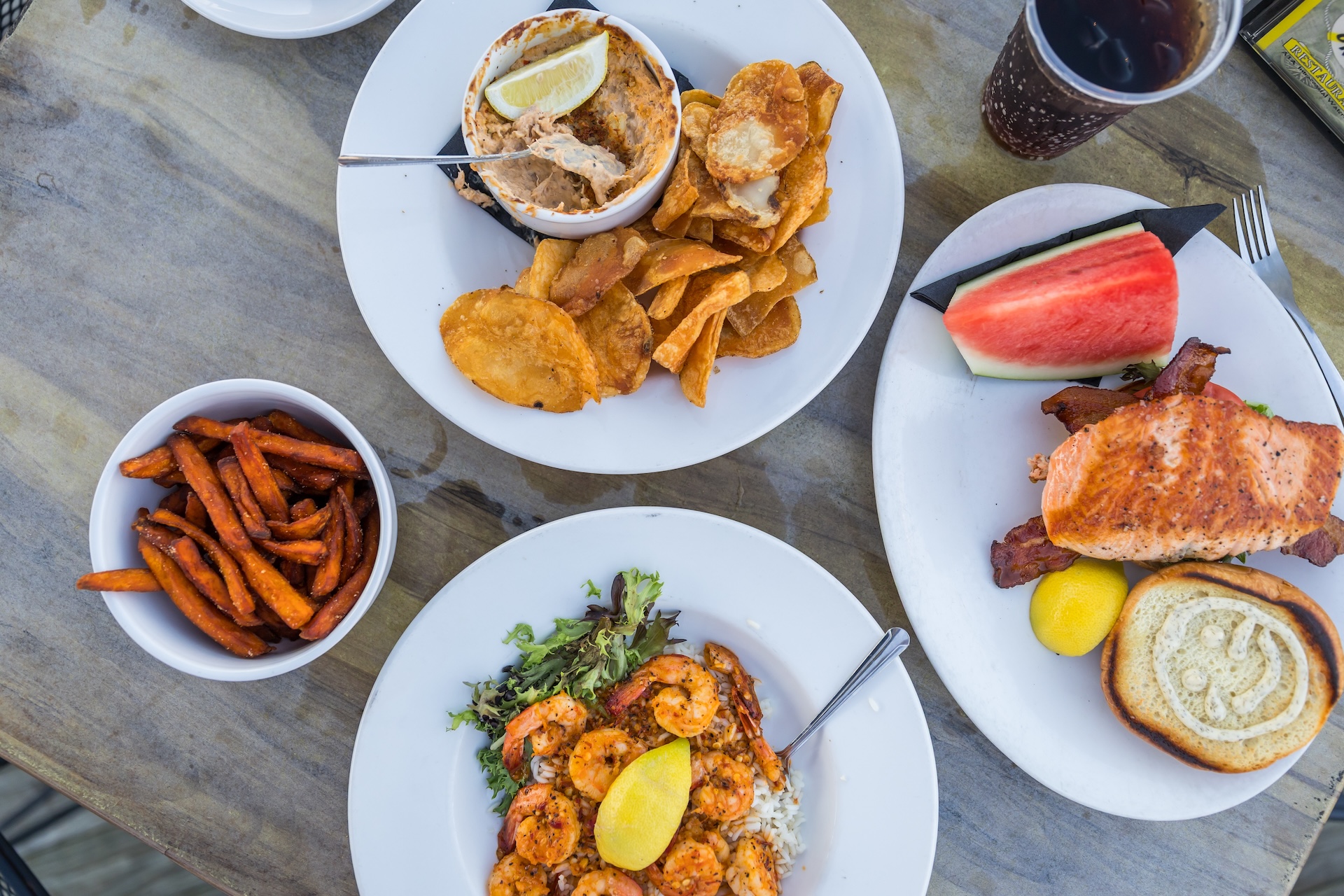 Four plates of food on a wooden table, featuring shrimp with rice and greens, sweet potato fries, a sandwich with bacon and watermelon, and a bowl of dip with thick-cut chips. There is also a drink with ice.