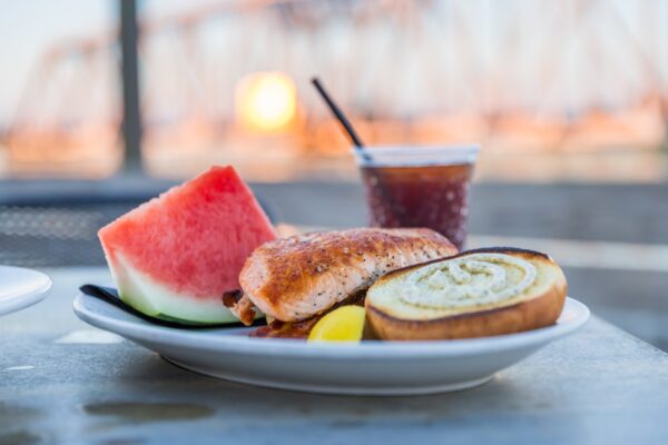 A plate with grilled salmon, a slice of watermelon, a lemon wedge, and a slice of bread sits on a table. A glass of dark iced beverage with a straw is behind the plate. A blurred bridge is seen in the background.