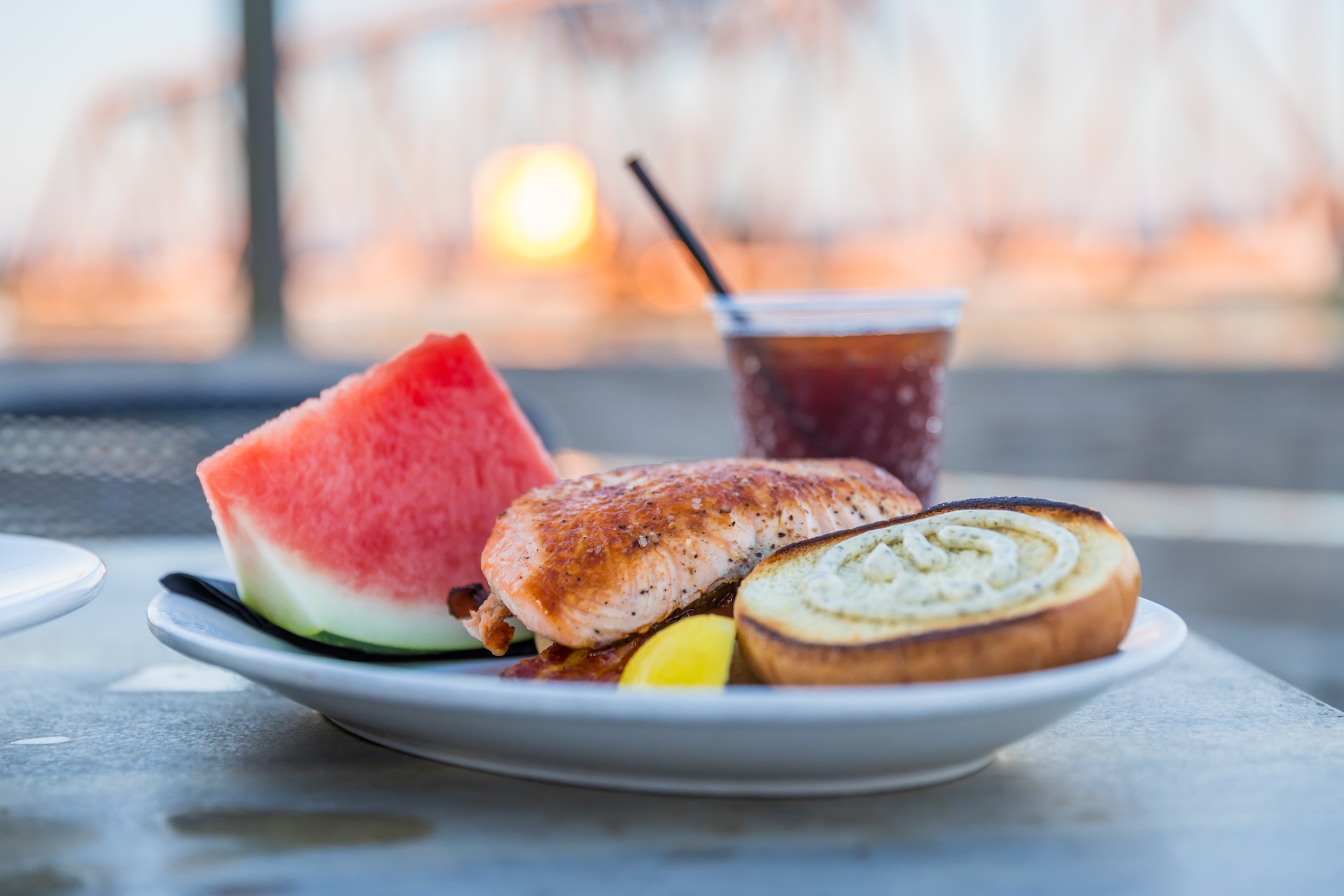 A plate with grilled salmon, a slice of watermelon, a lemon wedge, and a slice of bread sits on a table. A glass of dark iced beverage with a straw is behind the plate. A blurred bridge is seen in the background.