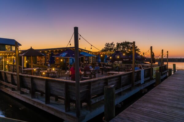 Outdoor dining area on a wooden deck by the water at sunset, with string lights, umbrellas, and people sitting at tables. The sky is vibrant with hues of orange and blue, creating a relaxed, inviting atmosphere.