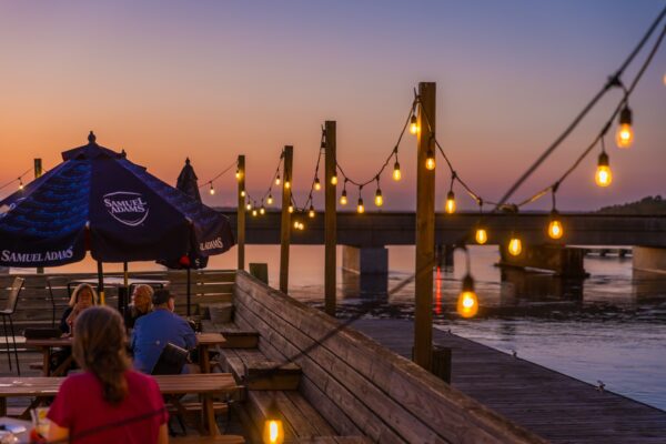 People sit at outdoor wooden tables under string lights and a blue umbrella by the water at sunset, with a bridge visible in the background and a warm, colorful sky.