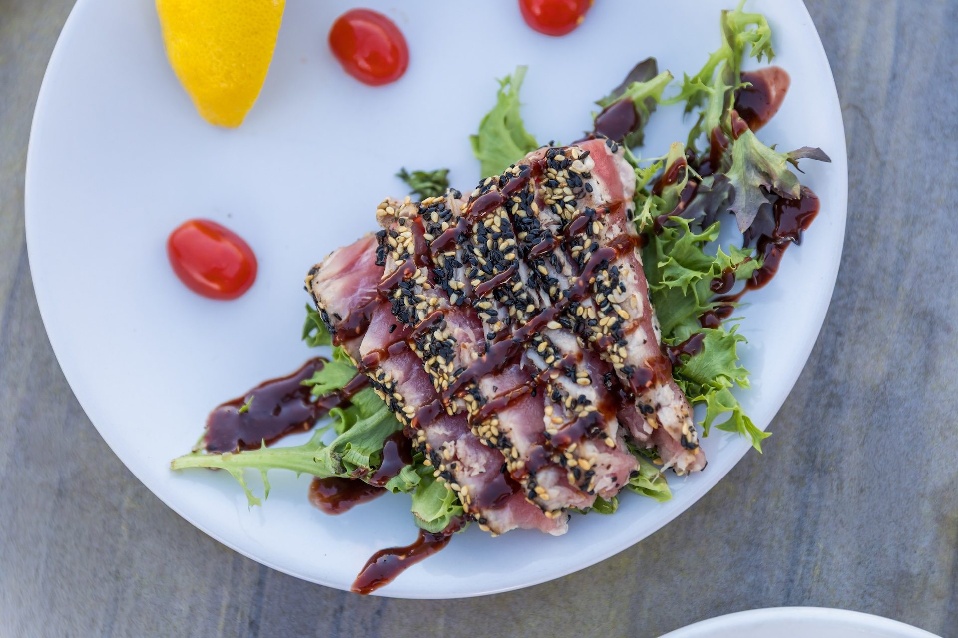 A plate with sesame-crusted seared tuna slices on mixed greens, drizzled with dark sauce. Cherry tomatoes and a lemon wedge are on the plate.