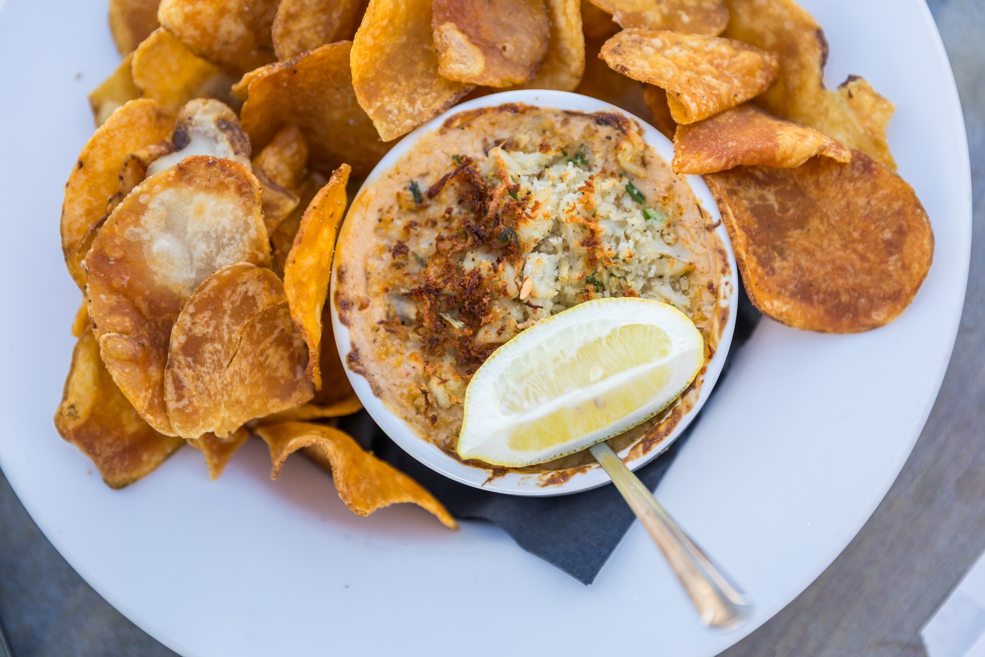 A white plate with crispy potato chips and a bowl of creamy dip, garnished with herbs, spices, and a lemon wedge. A spoon is resting in the dip.