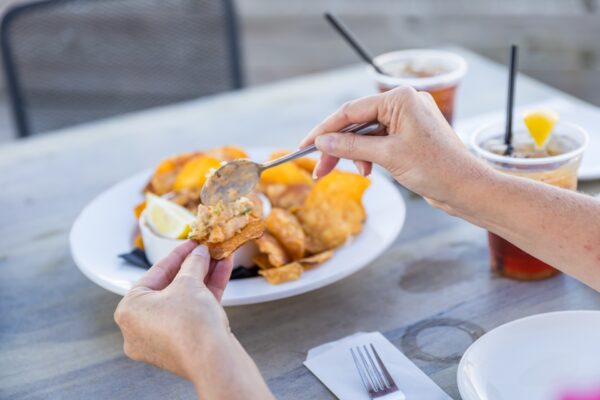 A person spreads a seafood dip onto a chip with a spoon, with a plate of chips, lemon wedges, and dip on the table, along with two iced drinks in plastic cups.
