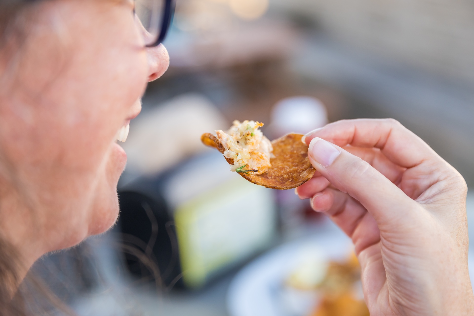 A person smiling as they hold a round cracker topped with a creamy dip close to their mouth, preparing to take a bite. The background is blurred, focusing on the hand and snack.