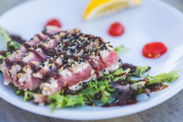 Slices of seared tuna coated with sesame seeds are served on leafy greens, drizzled with a dark sauce. Cherry tomato halves and a lemon wedge are on a white plate in the background.