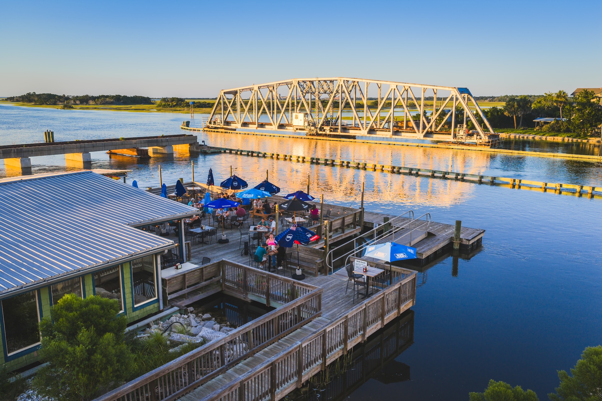 A waterfront restaurant with outdoor seating overlooks a calm river at sunset, with a white steel drawbridge in the background and patrons dining under blue umbrellas.