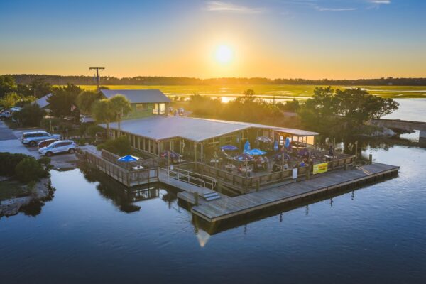 A waterfront restaurant with outdoor seating on a wooden deck, surrounded by water, with people dining as the sun sets over distant trees and marshland. Cars are parked nearby, and the sky is clear and bright.
