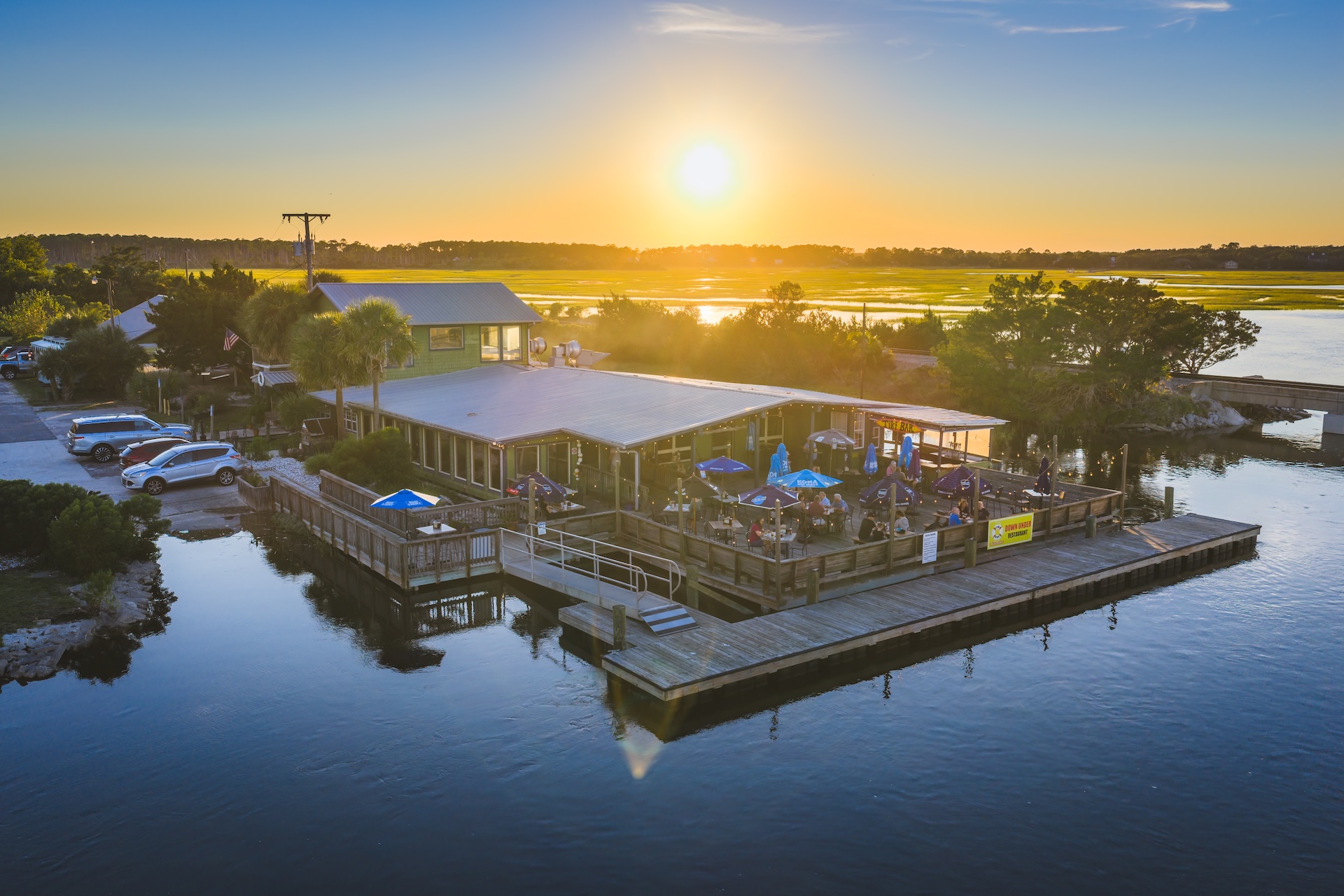 A waterfront restaurant with outdoor seating on a wooden deck, surrounded by water, with people dining as the sun sets over distant trees and marshland. Cars are parked nearby, and the sky is clear and bright.