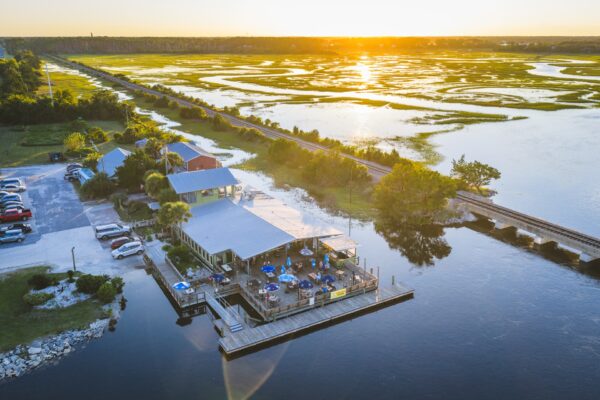 Aerial view of a waterfront restaurant with outdoor seating on a dock, surrounded by marshland at sunset. Several cars are parked nearby, and a railway line runs alongside the marsh.