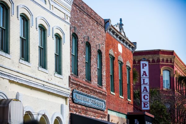 A row of historic brick buildings with tall windows and green shutters; a vertical red and white PALACE theater sign is visible on one building. The scene is bright and clear under a blue sky.