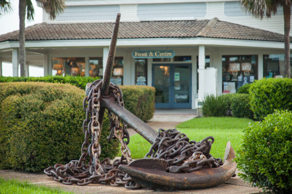 A large, rusted anchor with heavy chains is displayed on a manicured lawn in front of a shop called Front & Centre with glass doors and windows. Bushes and palm trees surround the anchor.