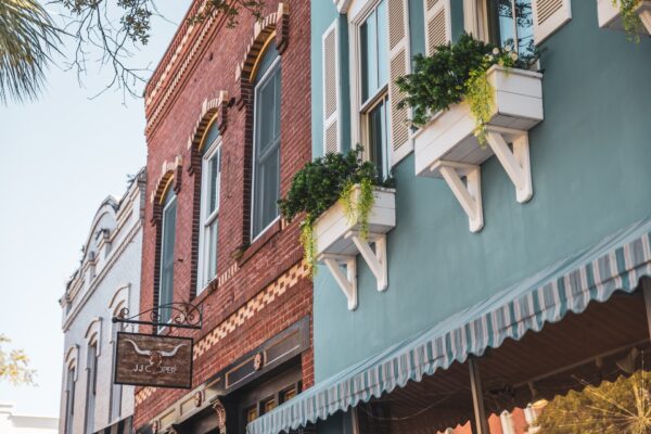 Colorful historic buildings with decorative windows, flower boxes, and an awning line a street. A metal shop sign hangs from the red brick building, and green plants spill from white window boxes on the blue building.