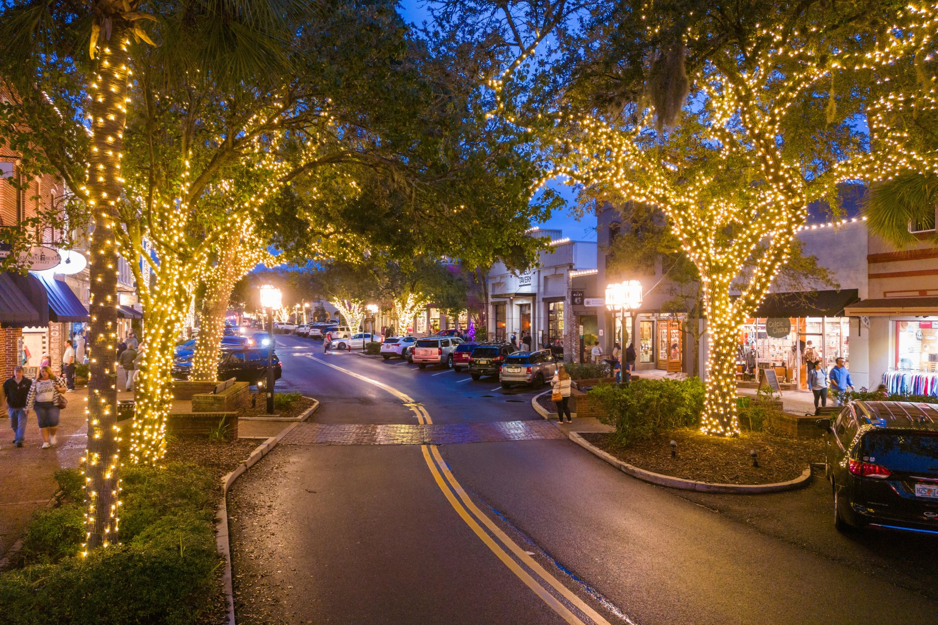 A charming downtown street at dusk with trees wrapped in twinkling lights, parked cars along the road, and people walking past shops and boutiques under a deep blue sky.