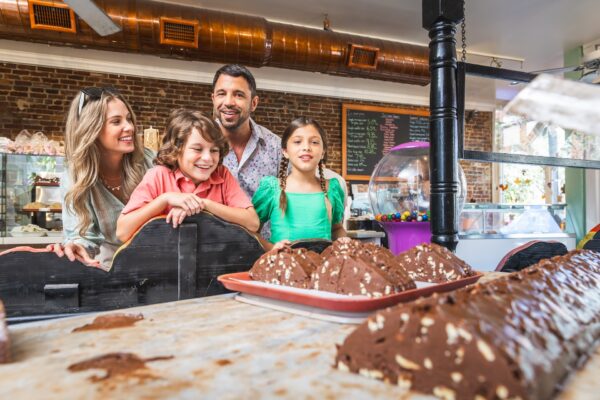 A smiling family of four admires a display of large chocolate treats inside a bakery or candy shop, with a chalkboard menu and gumball machine visible in the background.