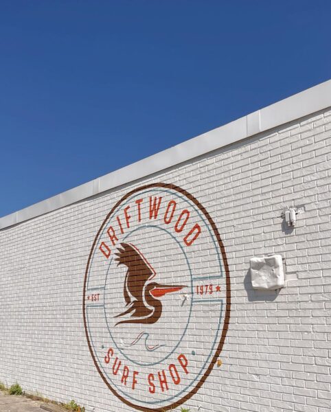 A white brick wall with a large round logo reading Driftwood Surf Shop and EST 1979, featuring a brown bird with outstretched wings, against a clear blue sky.