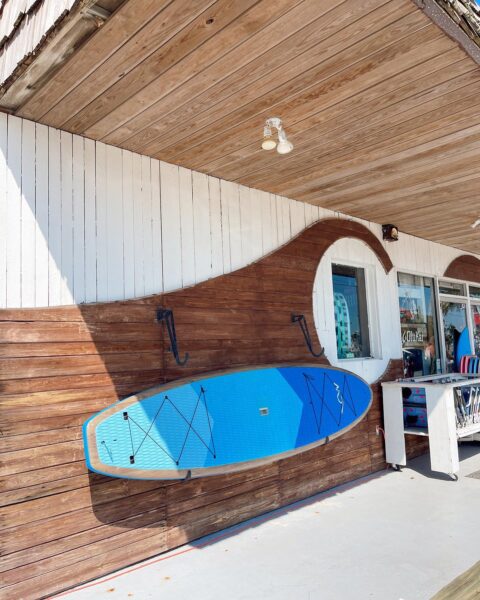 A blue paddleboard is mounted on a wall with a brown and white wave mural outside a shop. A window displays beach gear, and sunlight casts shadows on the board and wall.
