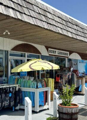 A surf shop with a Surf Lessons sign, colorful surfboards stacked under a yellow umbrella, clothing racks, and beach gear displayed outside under a shingled roof on a sunny day.
