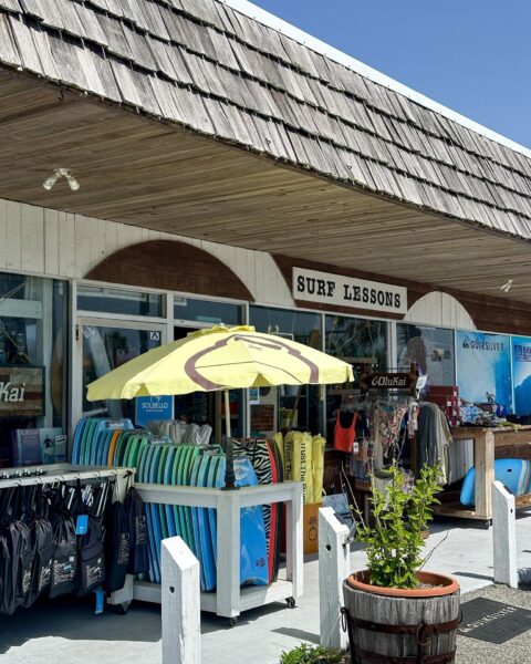 A surf shop with a Surf Lessons sign, colorful surfboards stacked under a yellow umbrella, clothing racks, and beach gear displayed outside under a shingled roof on a sunny day.