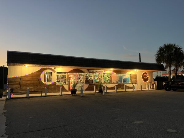A small roadside store called Sunny Shores is lit with string lights and decorated for Halloween at dusk. The parking lot is mostly empty, and a palm tree is visible to the right of the building.