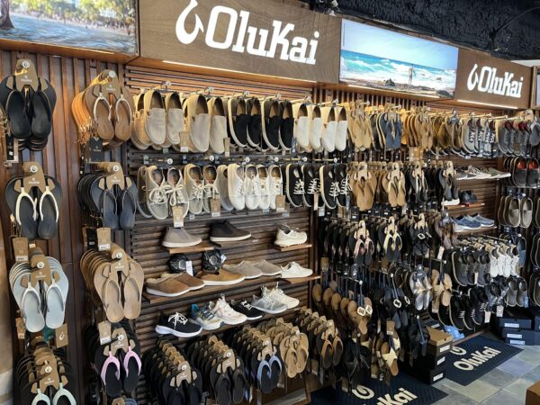 A retail display wall featuring numerous pairs of Olukai sandals and casual shoes organized on racks, with brand signage at the top.