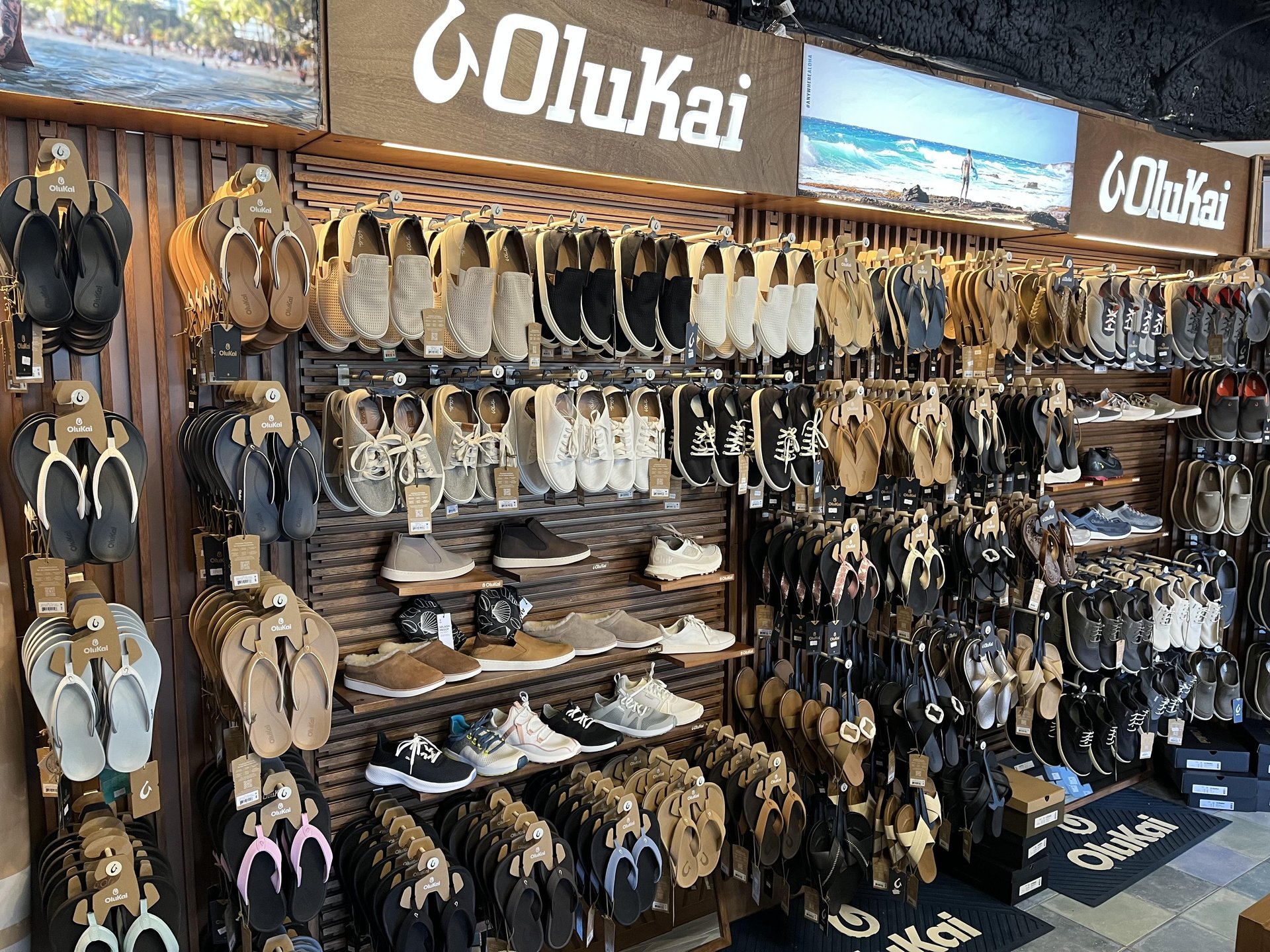 A retail display wall featuring numerous pairs of Olukai sandals and casual shoes organized on racks, with brand signage at the top.