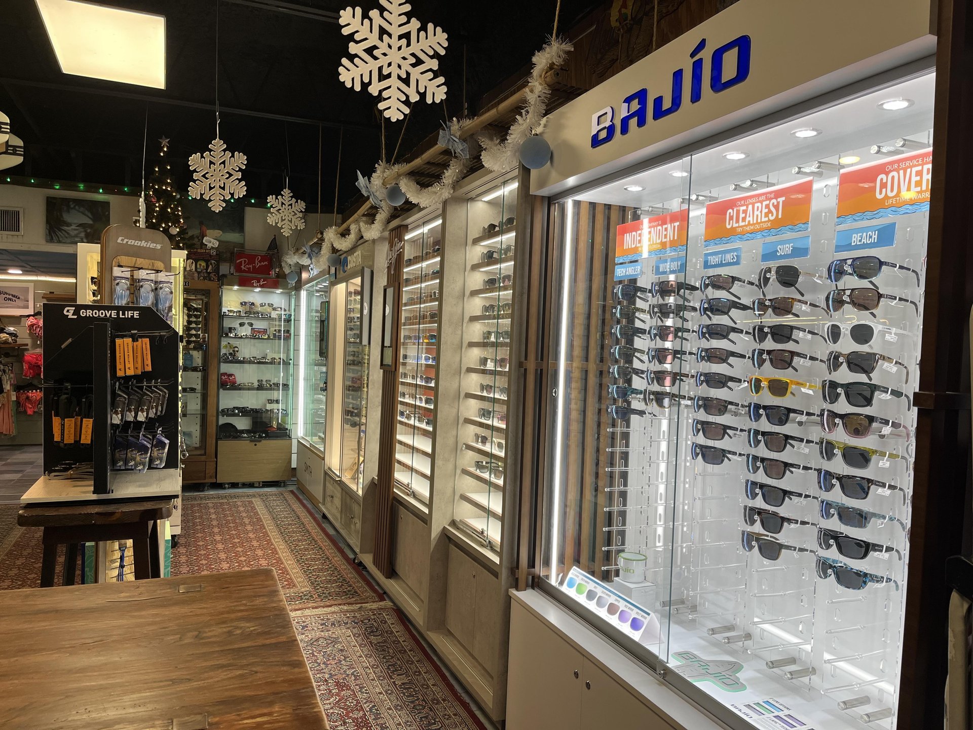 Display cases filled with rows of sunglasses inside a retail store, featuring prominent Bajío branding and overhead snowflake decorations.