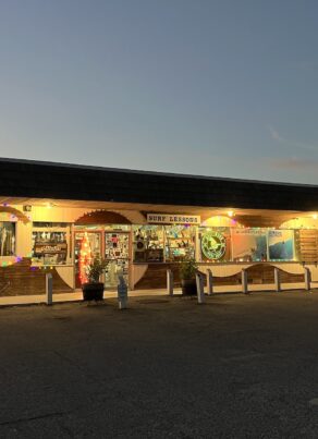 A surf shop with glowing string lights and colorful decorations on the exterior, photographed at dusk. A palm tree stands to the right, and a few cars are parked nearby. The store sign reads SUNNY SEASON.