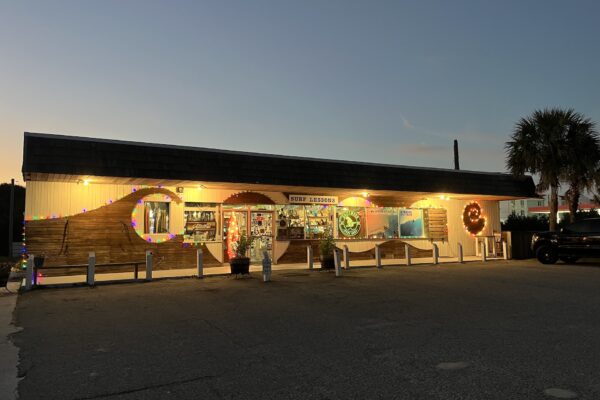 A surf shop with glowing string lights and colorful decorations on the exterior, photographed at dusk. A palm tree stands to the right, and a few cars are parked nearby. The store sign reads SUNNY SEASON.