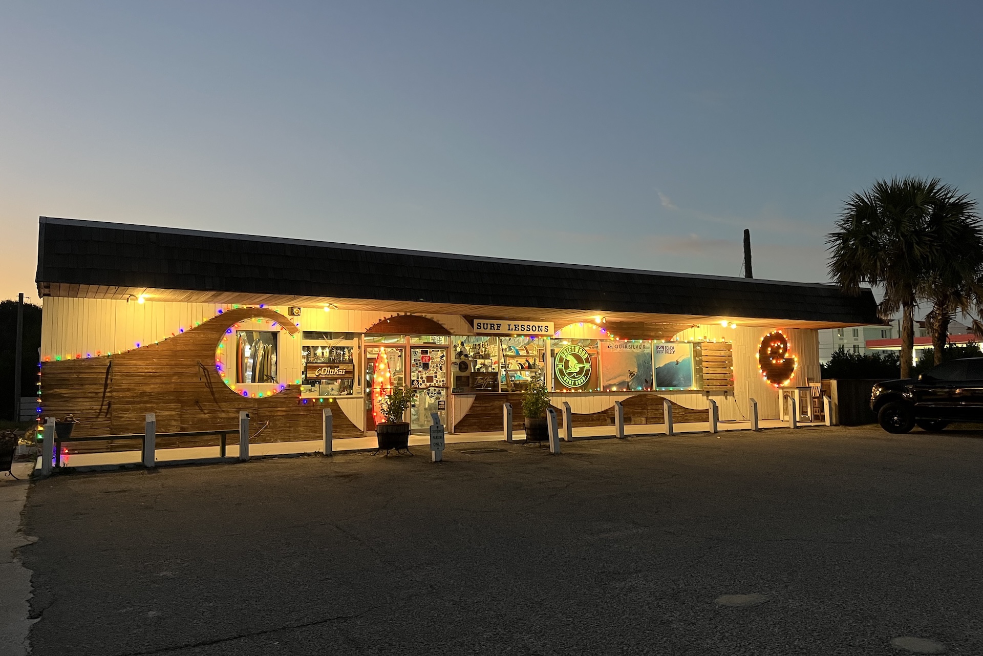 A surf shop with glowing string lights and colorful decorations on the exterior, photographed at dusk. A palm tree stands to the right, and a few cars are parked nearby. The store sign reads SUNNY SEASON.