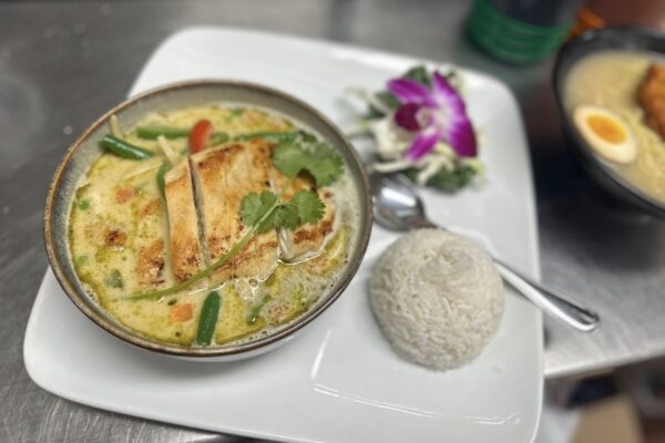 A bowl of Thai green curry with grilled chicken, vegetables, and cilantro garnish, served with a mound of white rice and a small side salad with an edible purple orchid on a white plate. A spoon is placed beside the rice.