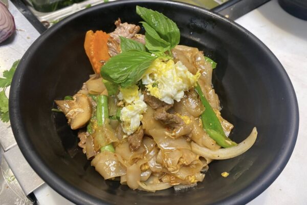 A black bowl filled with stir-fried wide rice noodles, beef, egg, onions, carrots, mushrooms, green vegetables, and fresh basil leaves on top.