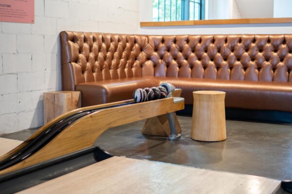 A cozy lounge area with a tufted brown leather sofa, two wooden stools, and a vintage mini bowling lane with several small bowling balls on the ball return.
