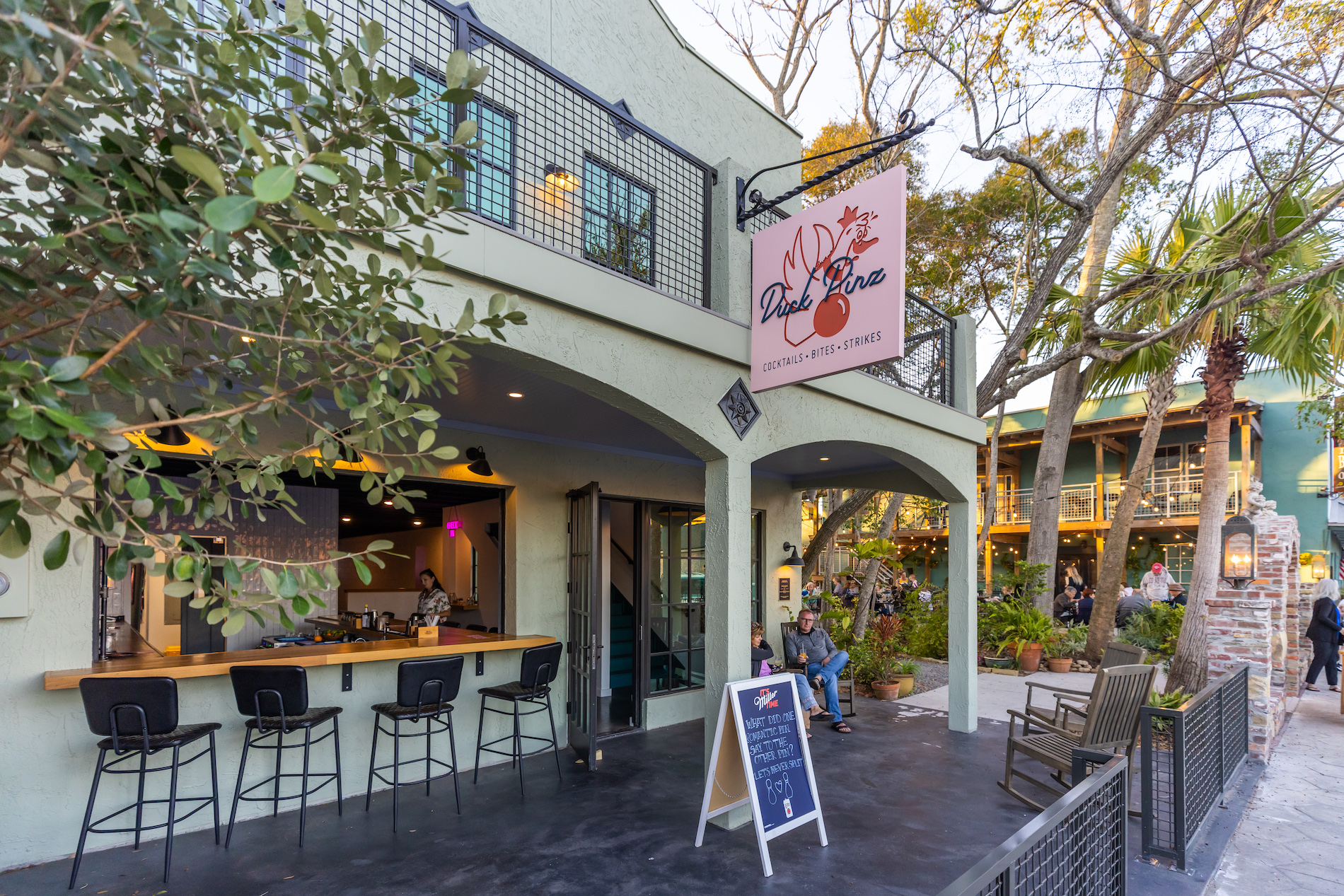 Outdoor view of a modern cafe with a covered patio, bar seating, and a large pink sign reading Duck & Son. People are sitting outside, and trees surround the building, creating a welcoming atmosphere.