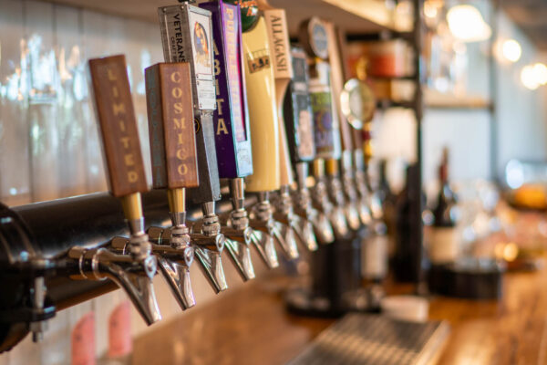 Close-up of several beer taps in a row at a bar, each with a different colorful handle and label. Glassware and bottles are visible on shelves in the background, and the bar has a warm, inviting atmosphere.