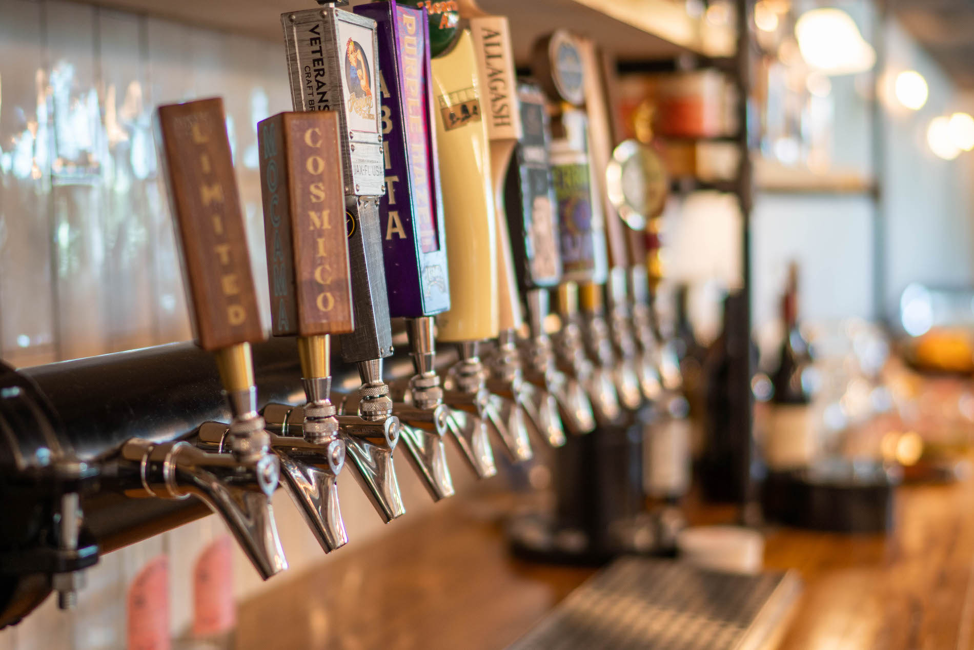 Close-up of several beer taps in a row at a bar, each with a different colorful handle and label. Glassware and bottles are visible on shelves in the background, and the bar has a warm, inviting atmosphere.
